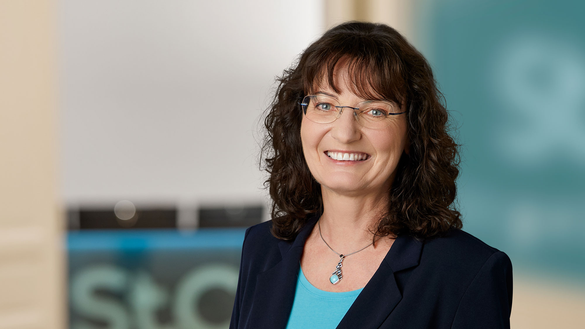 Portrait einer l&auml;chelnden Frau mit Brille und lockigem Haar, unscharfer Hintergrund mit dem Logo 'St.'