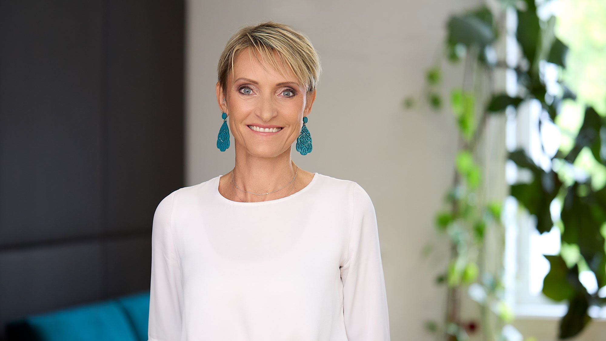 Eine Frau mit kurzem blondem Haar und blauer Ohrringe steht l&auml;chelnd in einem Raum mit Pflanzenhintergrund.