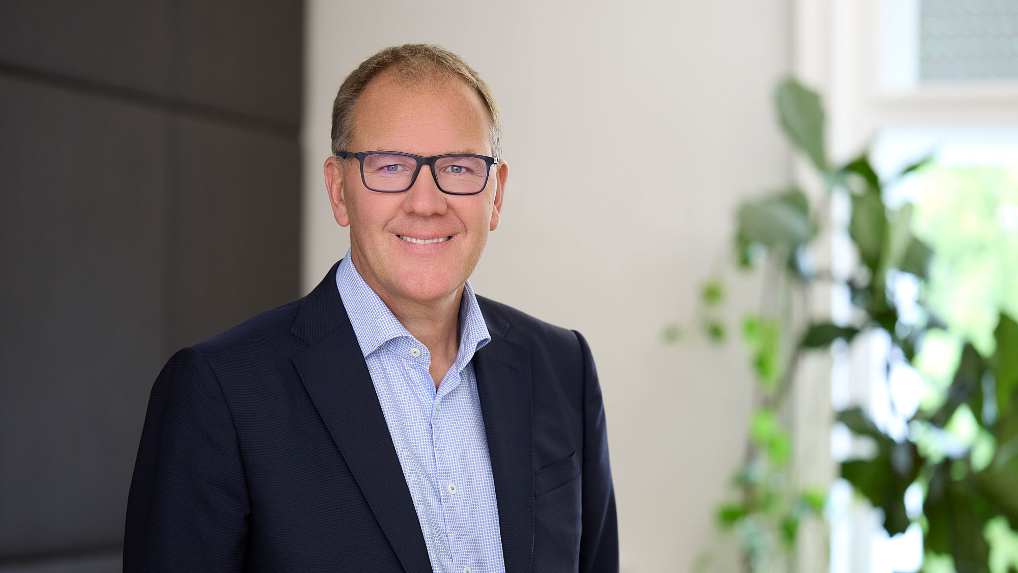 Ein l&auml;chelnder Mann mit Brille und Anzug vor einem verschwommenen Hintergrund mit Pflanzen.