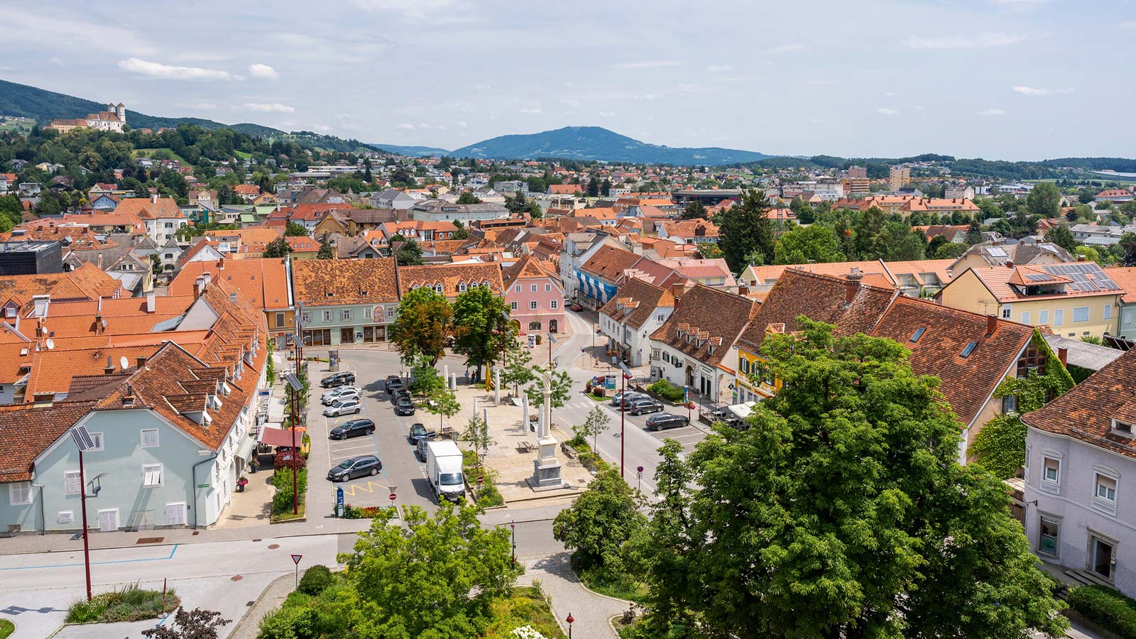 Luftaufnahme einer Stadt mit roten D&auml;chern, einem zentralen Platz und einer h&uuml;geligen Landschaft im Hintergrund.